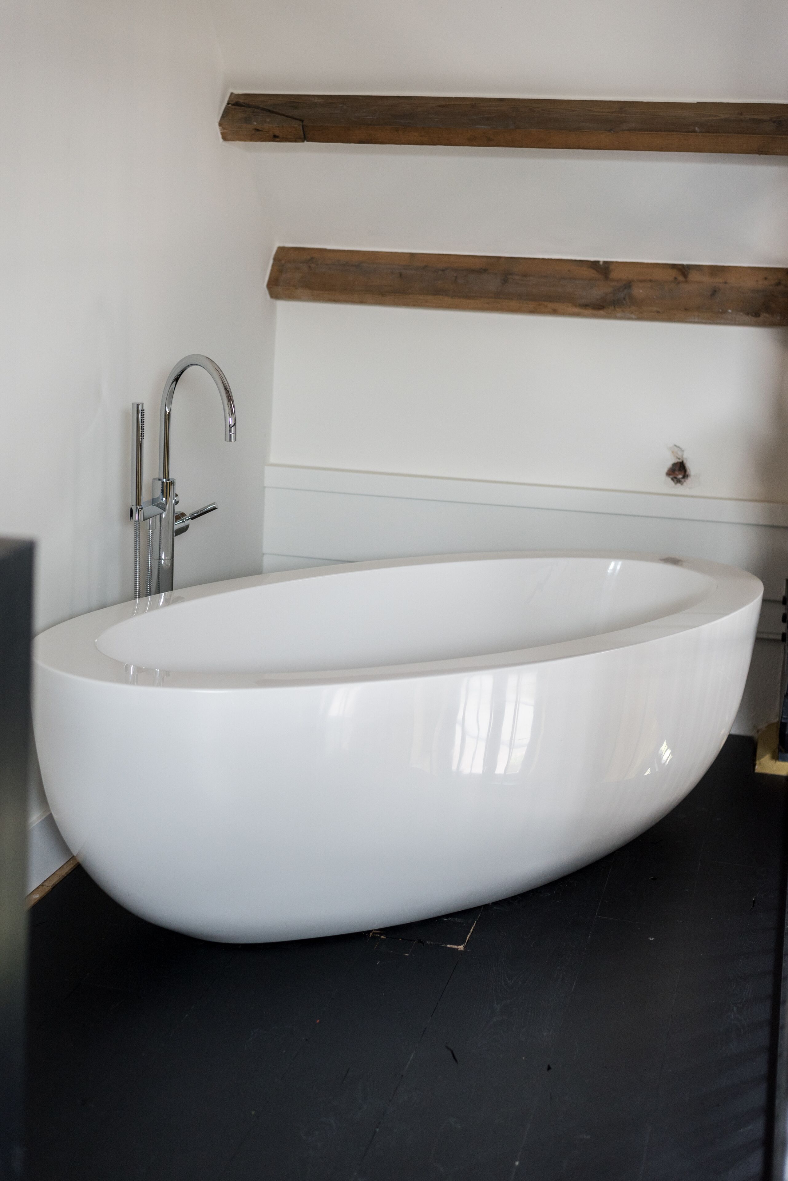 A modern bathroom with a freestanding bathtub, a chrome faucet, and a wooden beam above the tub.
