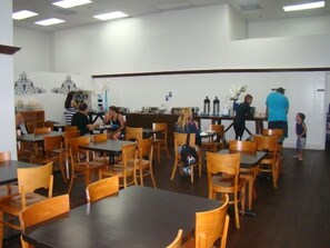 A restaurant with wooden tables and chairs, a counter with a person serving, and a child standing near the entrance.