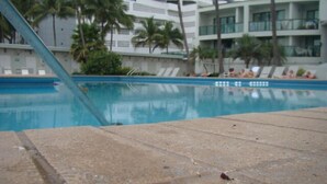 A swimming pool with a clear blue water surface, surrounded by palm trees and a modern building with balconies in the background.
