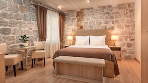 Een slaapkamer met een stenen muur, een bed met een houten hoofdeinde, een bankje, een kleine tafel met een plant, en twee bedlampen.