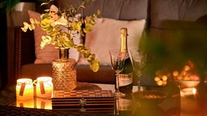 Een knus interieur met een rieten tafeltje, kaarsen, een fles champagne en een vaas met bloemen.
