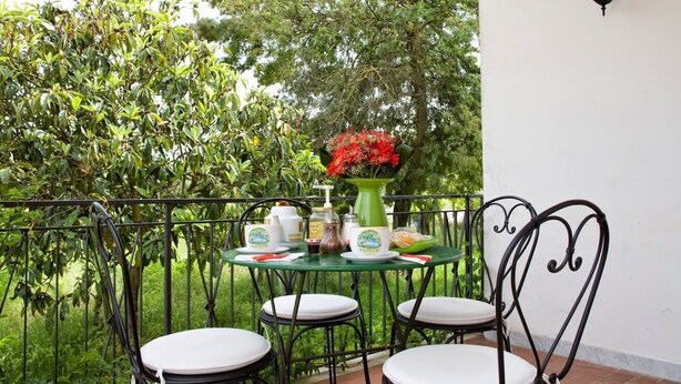 Una pequeña mesa al aire libre con sillas, un jarrón de flores rojas y un farol de pared.