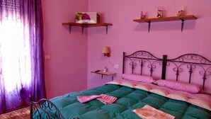 Un dormitorio con colcha verde acolchada, estructura de cama metálica y almohadas rosadas. Hay dos repisas de madera sobre la cama con adornos.