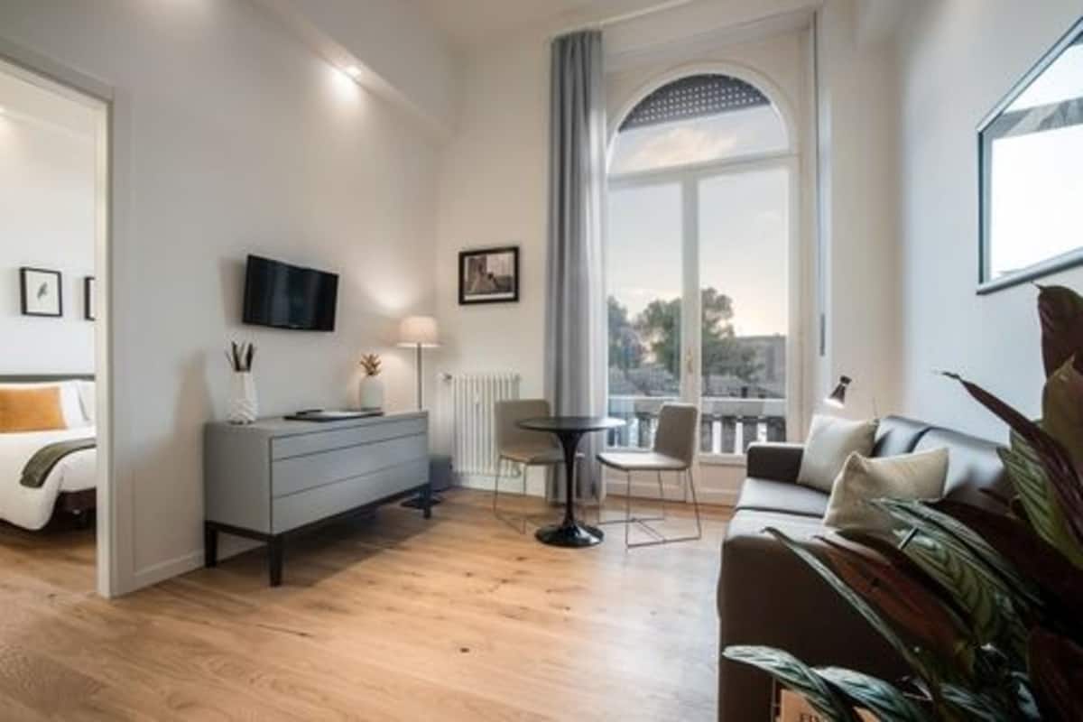 A modern living room with a sofa, a coffee table, and a TV mounted on the wall. There is a window with a view of the outdoors.