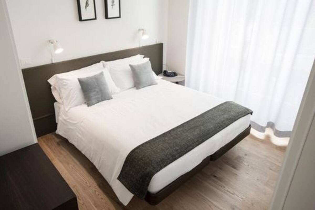 A neatly made bed with white bedding and a dark headboard, a bedside table with a lamp, and a window with sheer curtains.