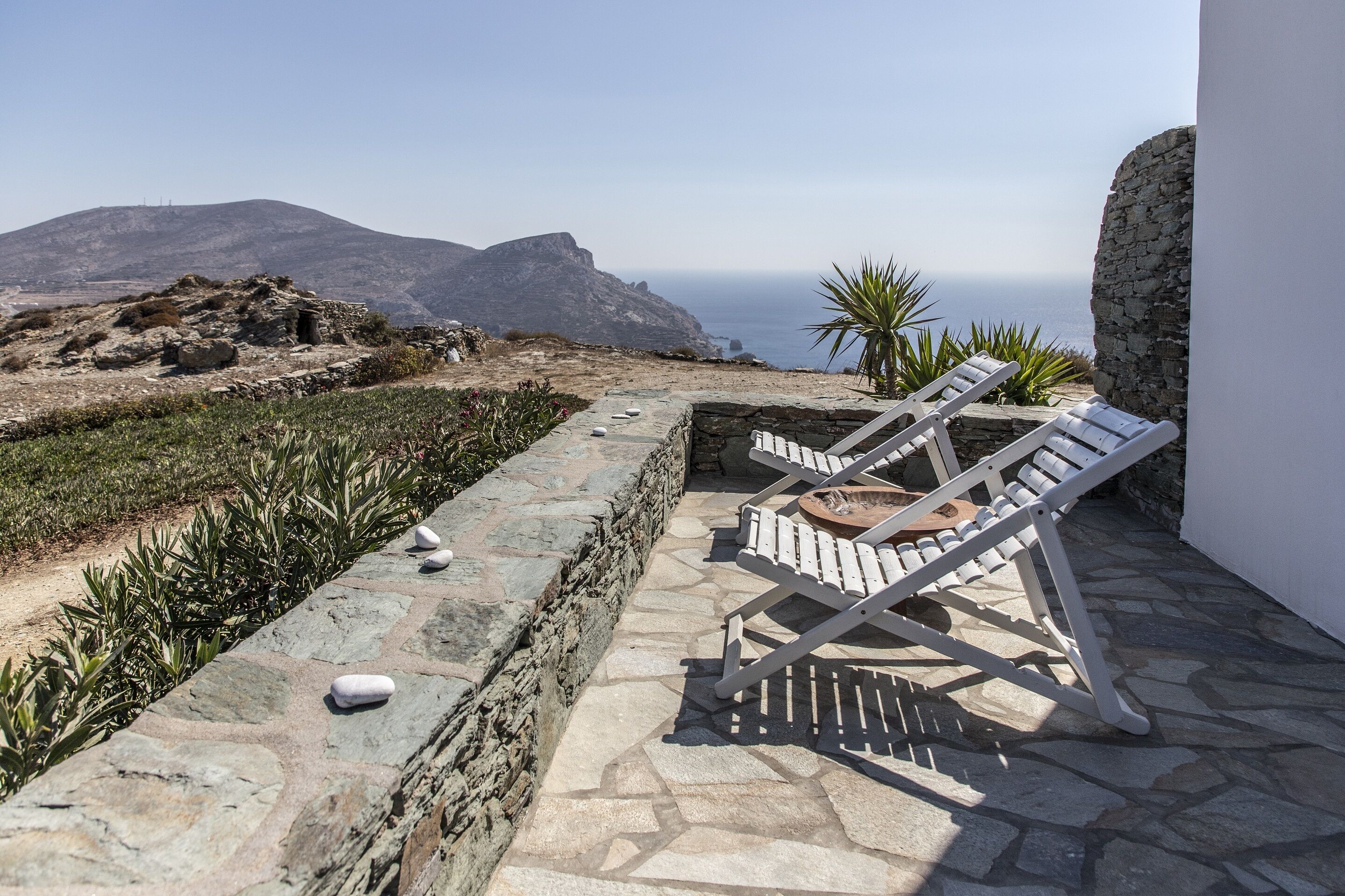 Un chemin de pierre mène à une terrasse où se trouvent des chaises longues blanches, d’où l’on peut admirer un paysage côtier.