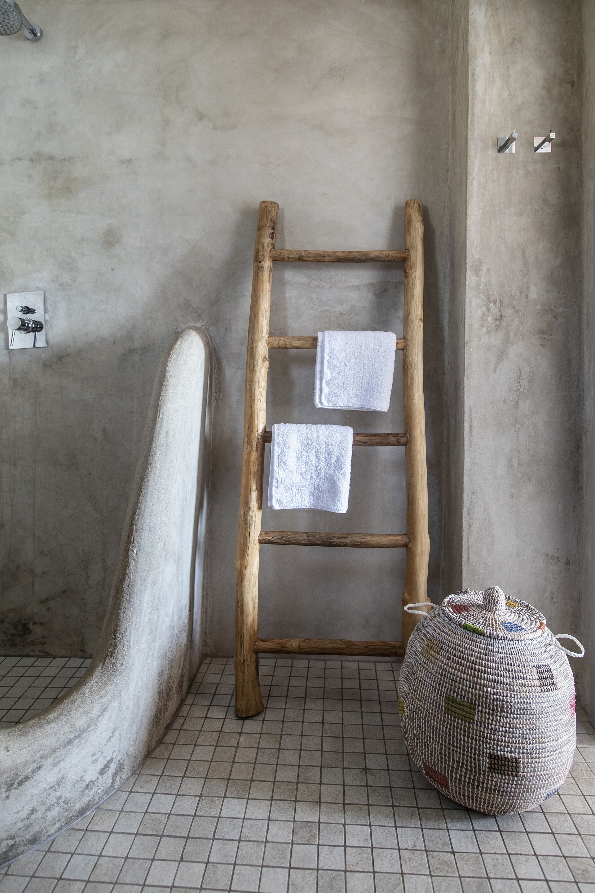Une salle de bain avec une échelle en bois, deux serviettes blanches, un pouf en fibres tressées et un sol carrelé.