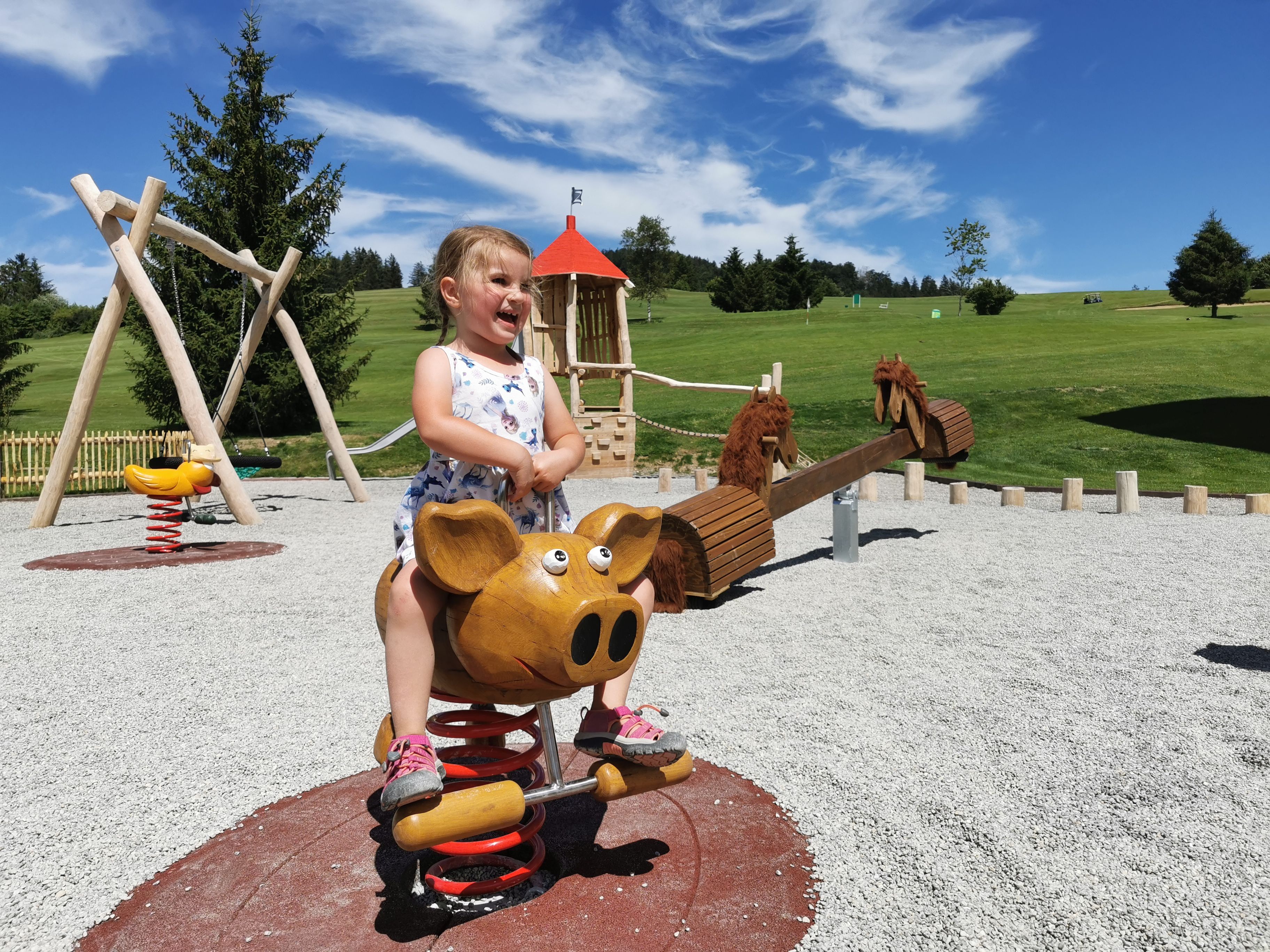A child on a playground ride with a wooden structure and a red-roofed playhouse in the background.