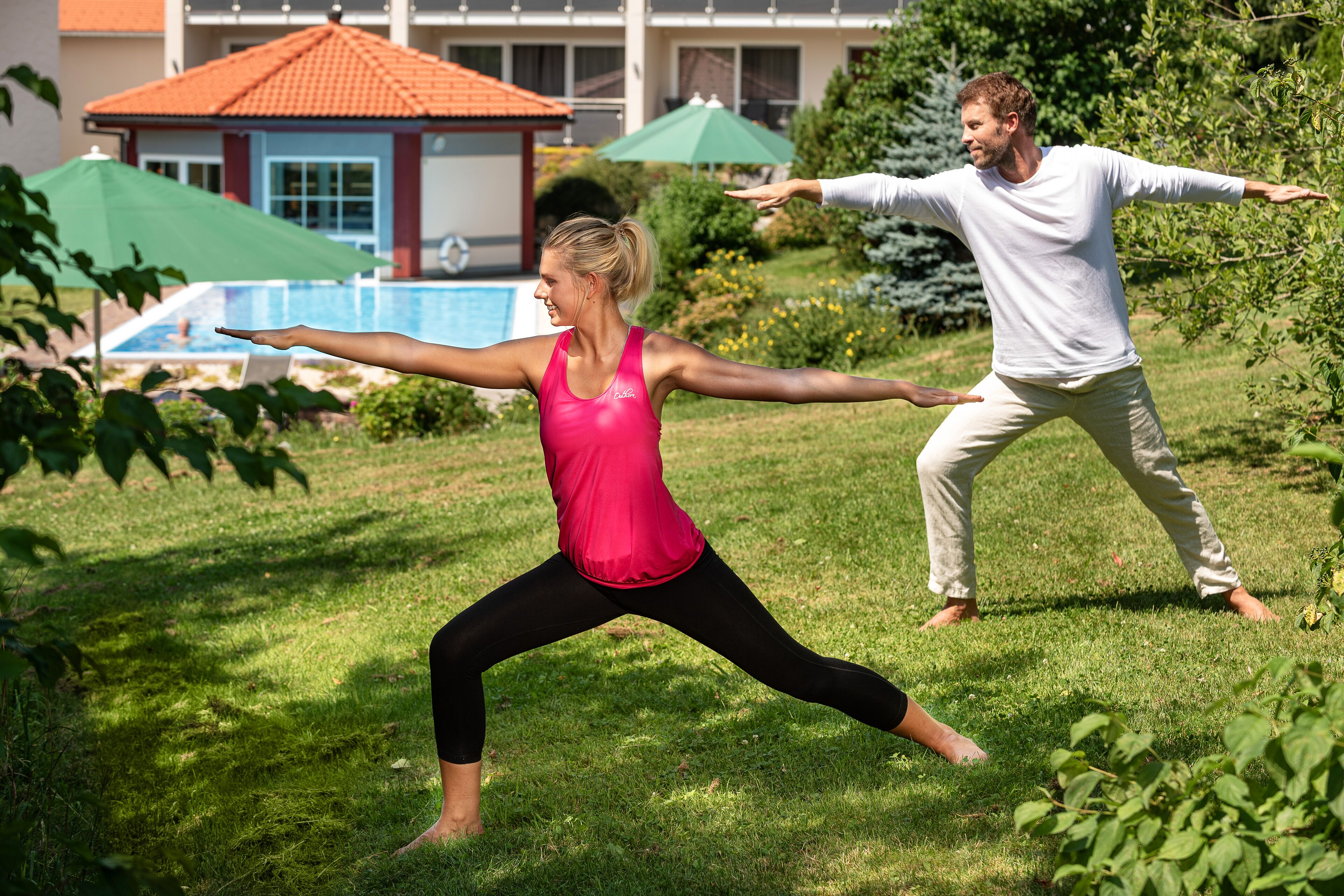 Two people practicing yoga outdoors in a grassy area with a pool and umbrellas in the background.