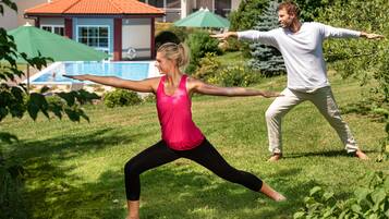 Two people practicing yoga outdoors in a grassy area with a pool and umbrellas in the background.