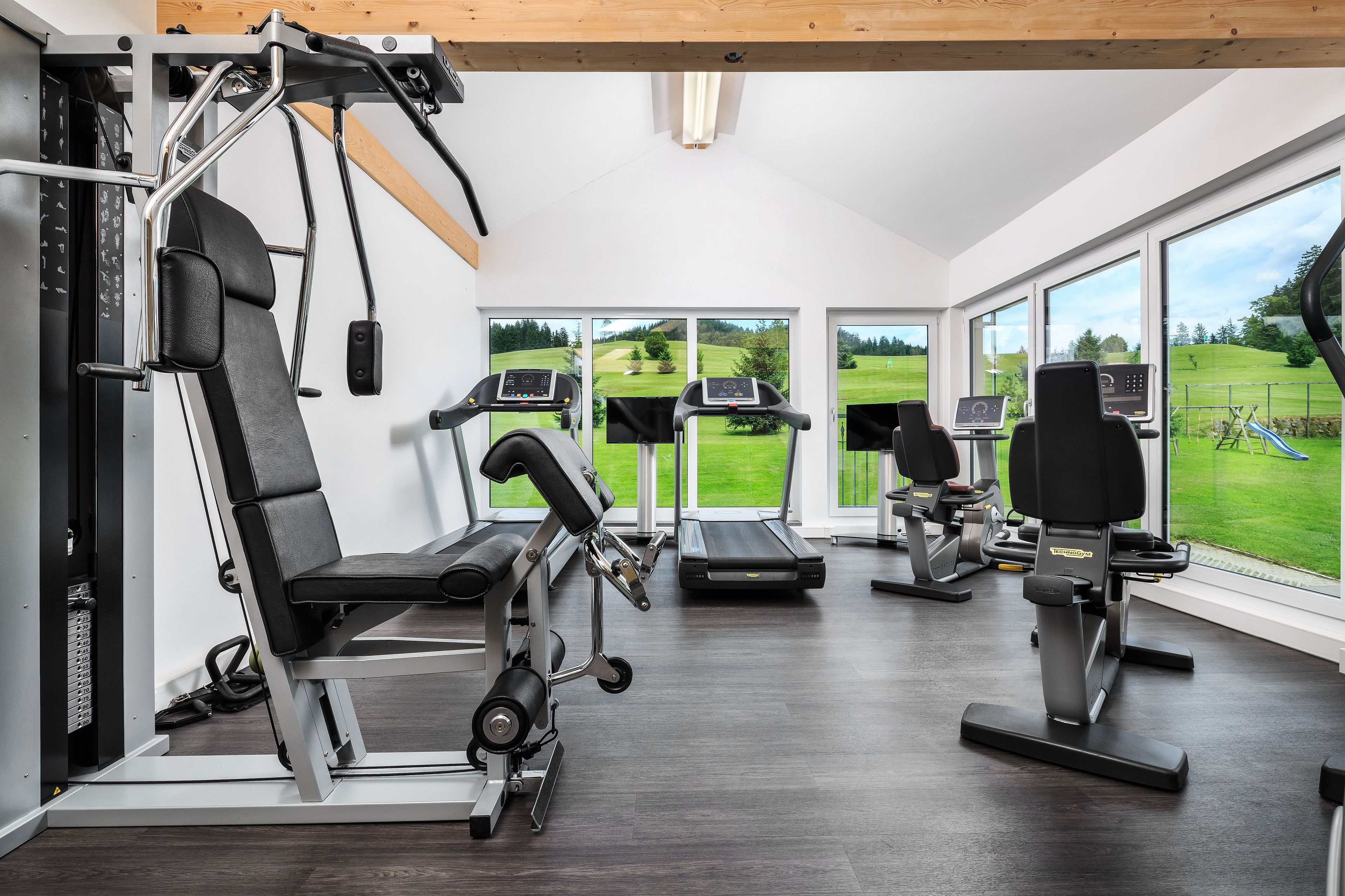 A gym with cardio machines, weight training equipment, and large windows offering views of a green landscape.