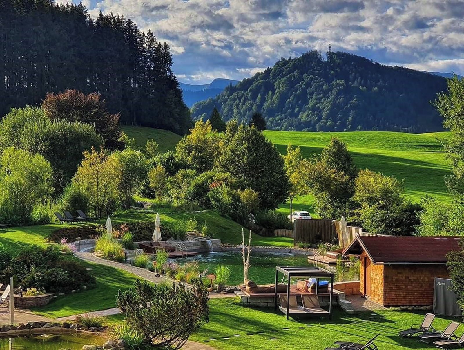 Ein üppiger Garten mit Teich, Sitzgelegenheiten im Freien und einem kleinen Gebäude, umgeben von Hügeln und Bäumen.