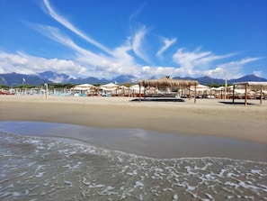 Ein Strand mit Sonnenschirmen und Liegestühlen, einem klaren Himmel und Bergen im Hintergrund.