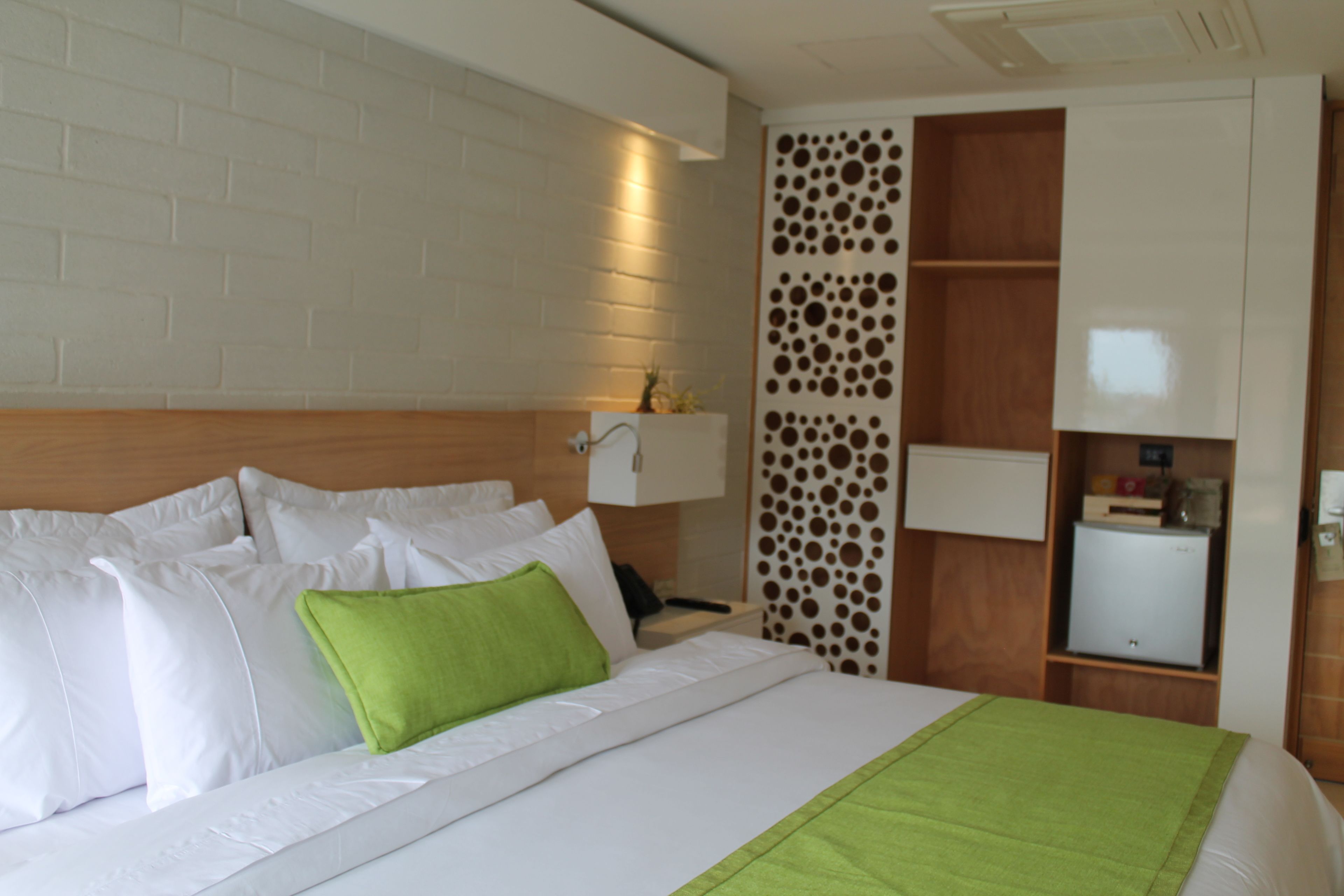 Een hotelkamer met een groot bed, een houten hoofdbord, een witte bakstenen muur, een decoratief paneel, een nachtkastje met een lamp en een kleine koelkast.