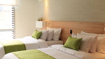 Habitación de hotel con dos camas, cada una con ropa de cama blanca y detalles verdes. Hay una planta pequeña en un estante sobre las camas, y una ventana con persianas a la izquierda.