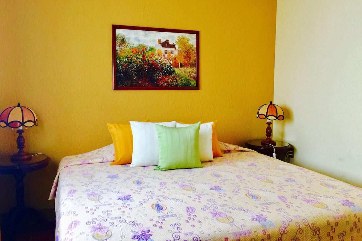 A bedroom with a bed, pillows, a painting, and two lamps.