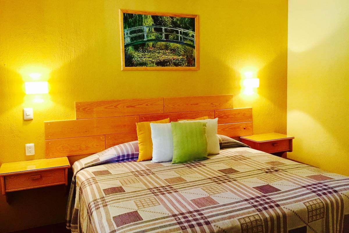 A room with a bed, wooden headboard, two bedside tables, and a painting of a bridge.