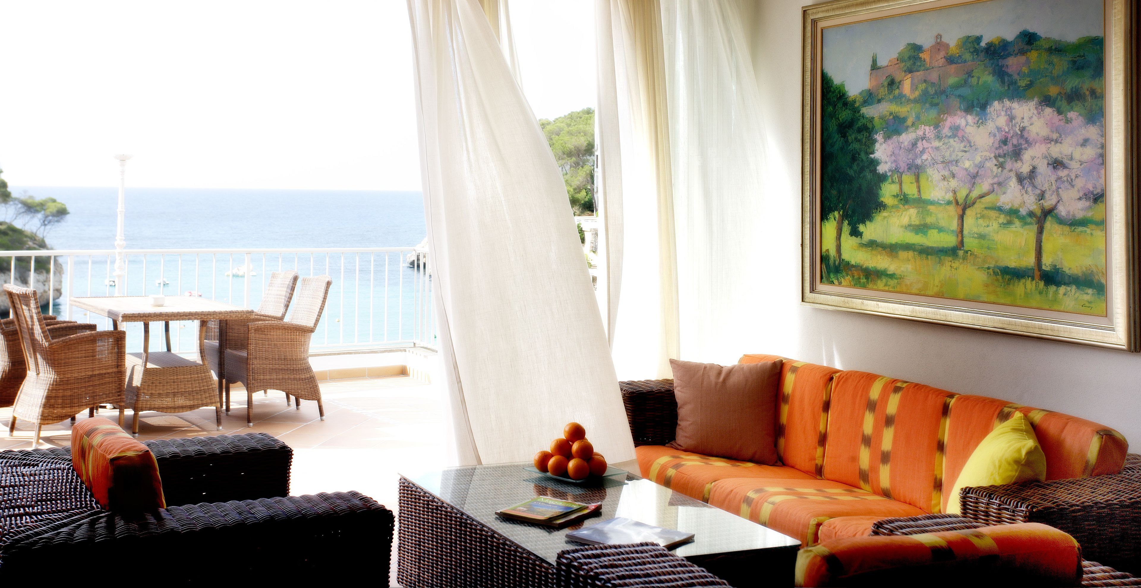 Un balcon agrémenté de meubles en osier, une table en verre avec des oranges et un tableau représentant des arbres.