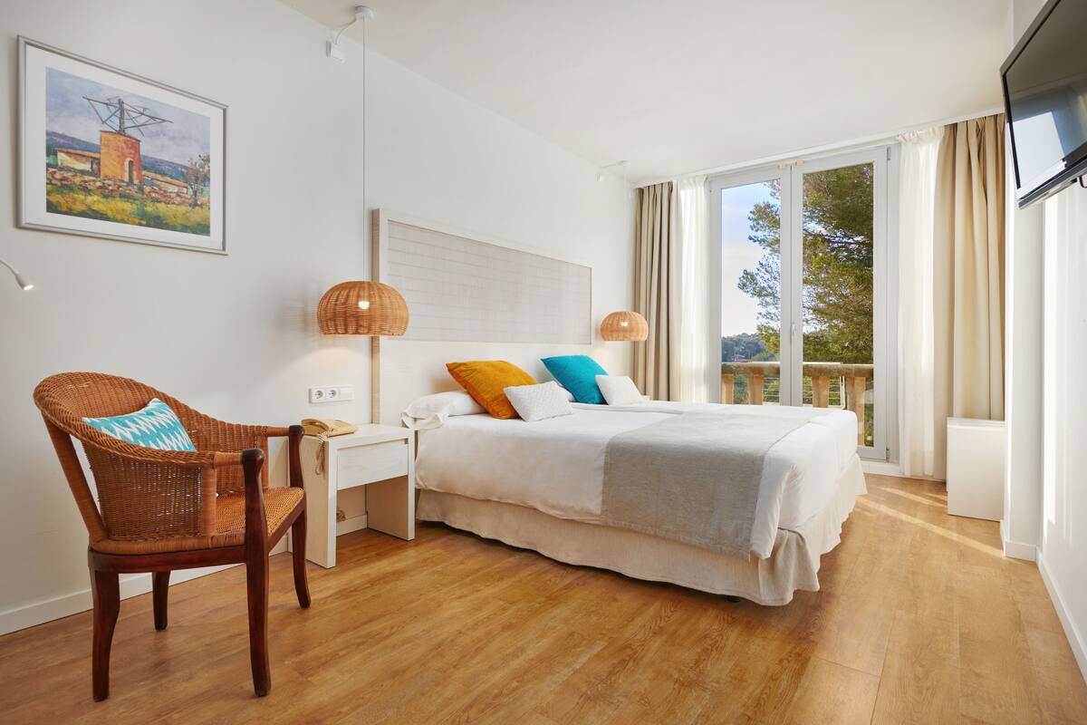 A bedroom with a bed, a wicker chair, a bedside table, a lamp, and a painting on the wall.