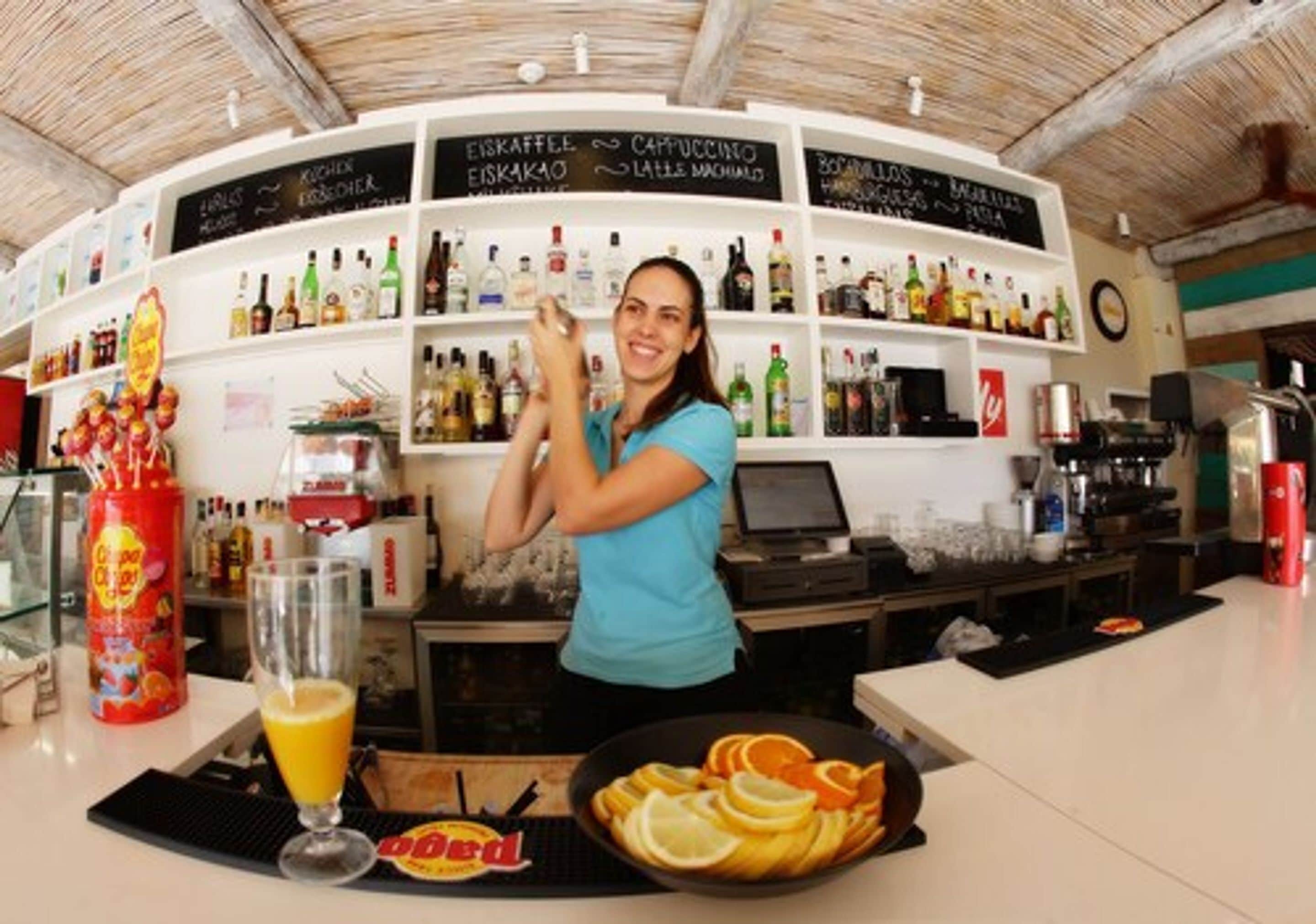 Una zona de bar con estantes llenos de botellas de licores, un vaso de zumo de naranja y un bol de rodajas de naranja.