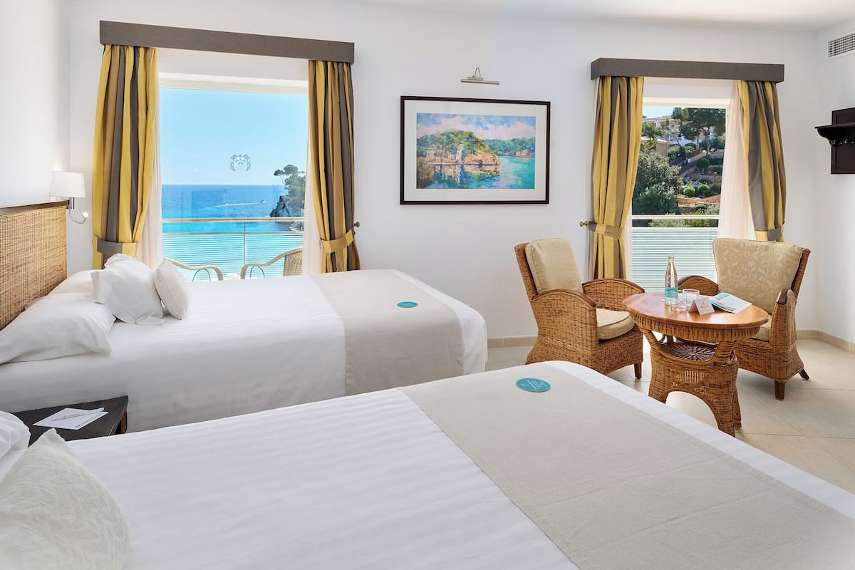 A hotel room with two beds, a dining table with chairs, and large windows with curtains.