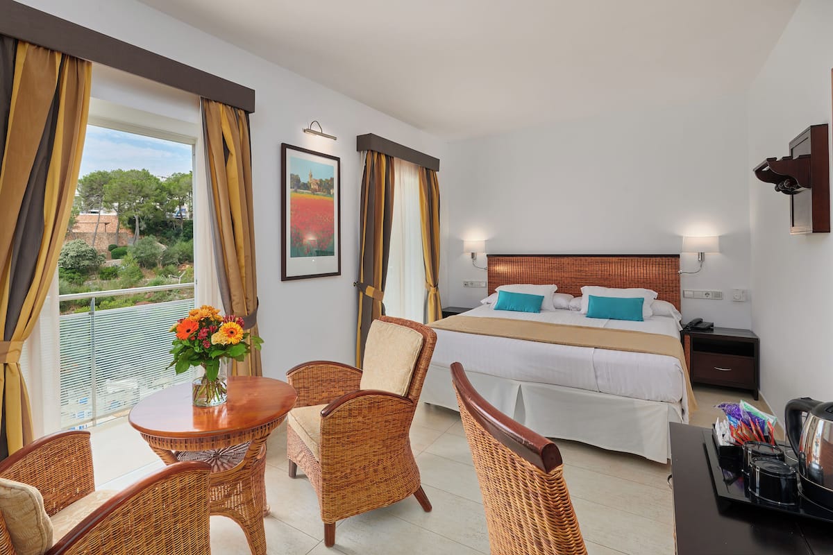 A hotel room with a bed, a dining area with wicker furniture, and a view of greenery outside.