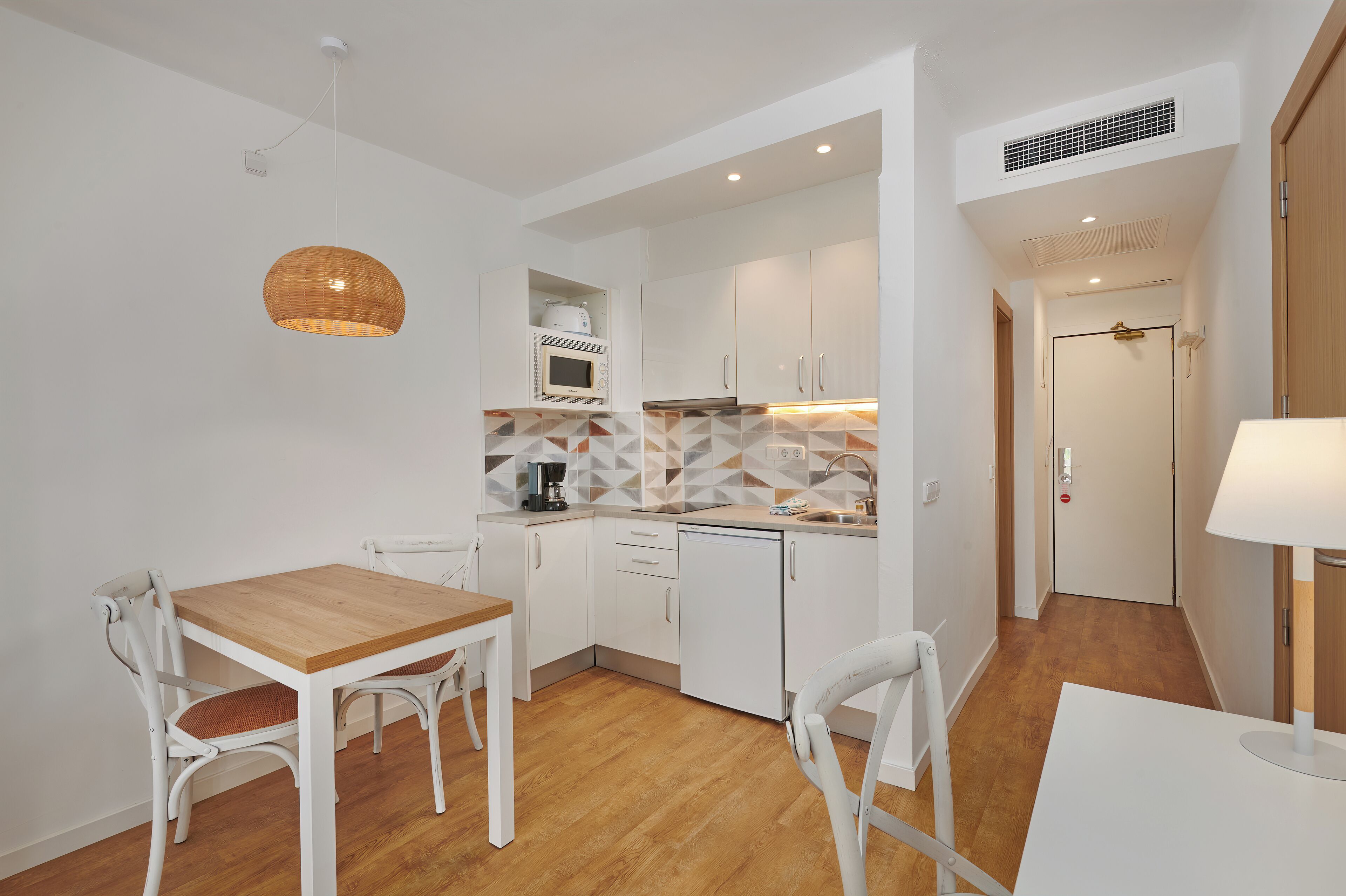 A compact kitchen with white cabinets, a microwave, and a coffee maker. There is a dining table with chairs and a hanging light fixture.