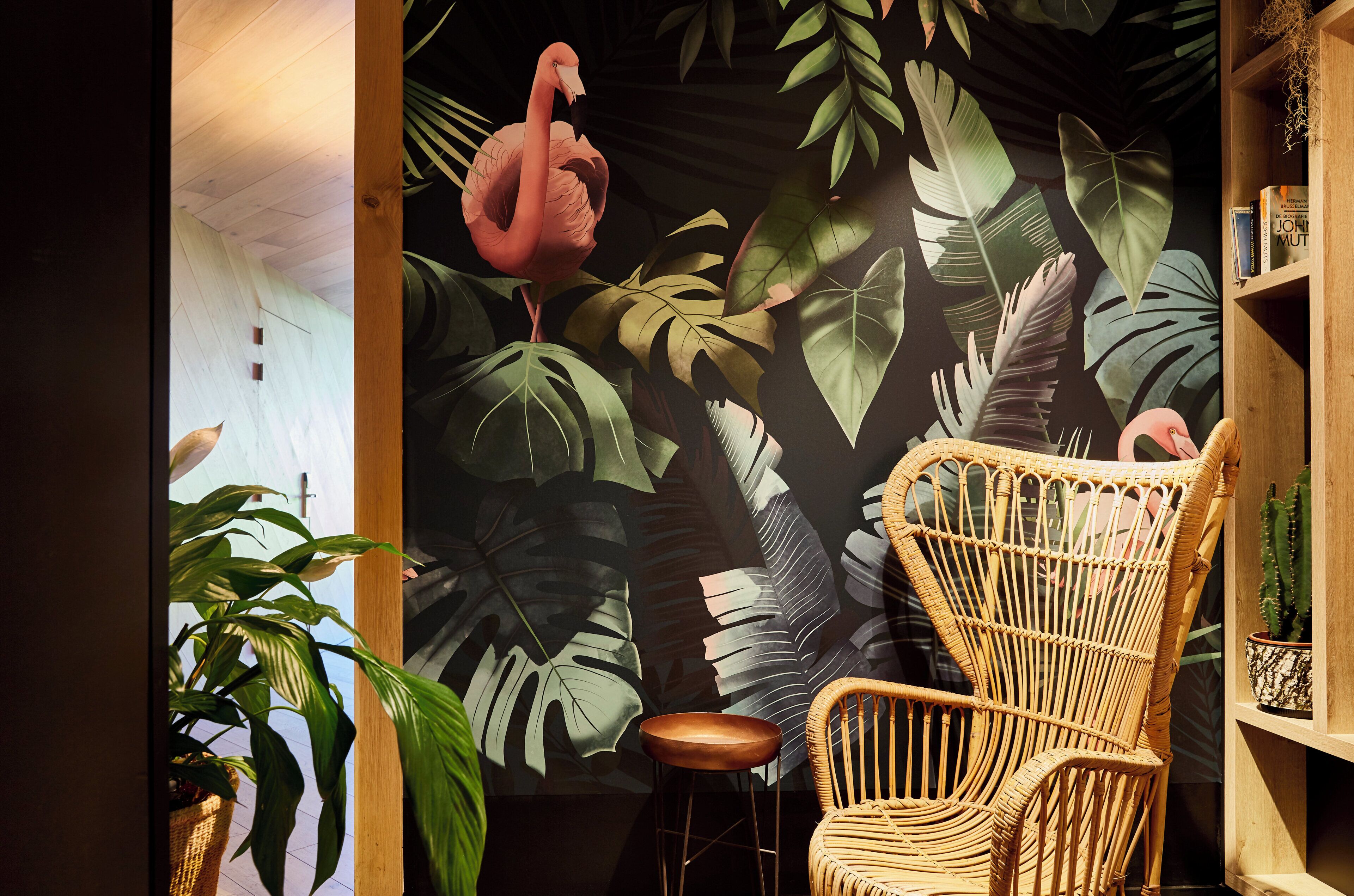 Una stanza con un murale di foglie tropicali e un fenicottero, una sedia in vimini e una libreria piena di libri.
