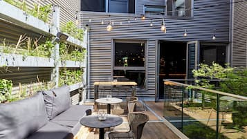 Un espace extérieur aménagé avec un canapé, des tables et des chaises, entouré de plantes et d’une rambarde.