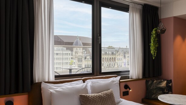 Une chambre d’hôtel avec un lit, une petite table, une plante suspendue et une vue sur des bâtiments historiques.