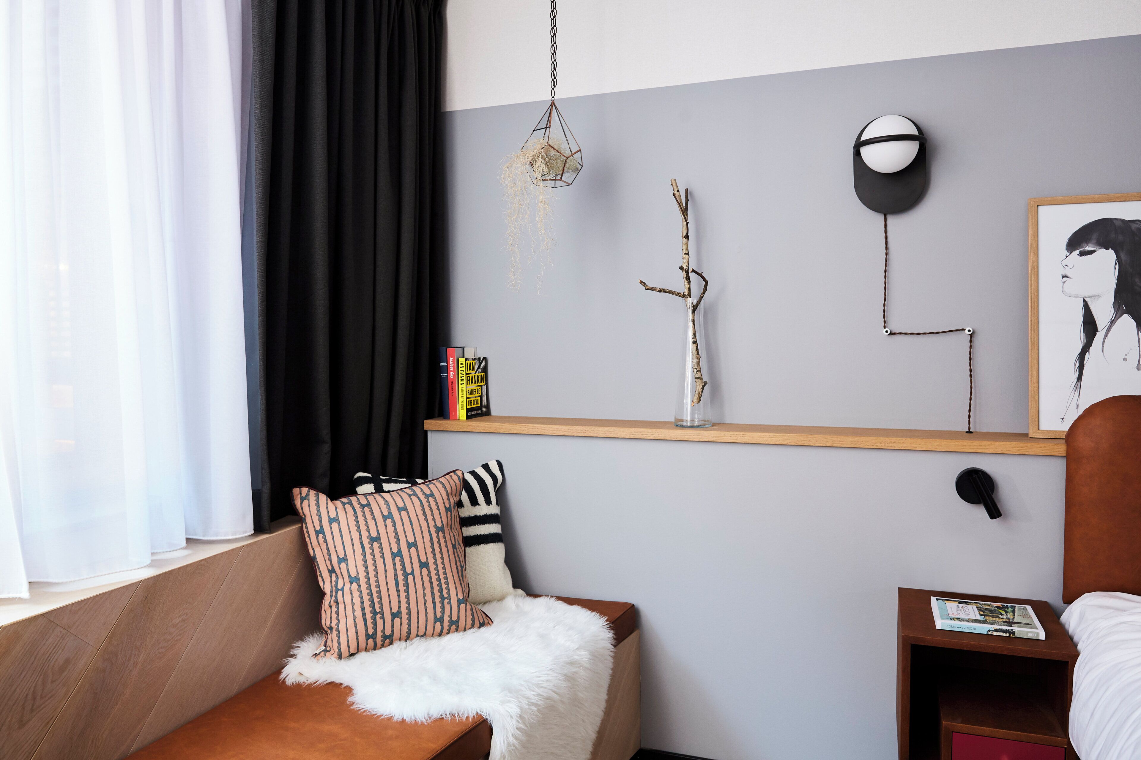 A modern bedroom with a wooden bed, a white furry throw, a black and white pillow, a shelf with books and decorative items, and a wall-mounted light fixture.