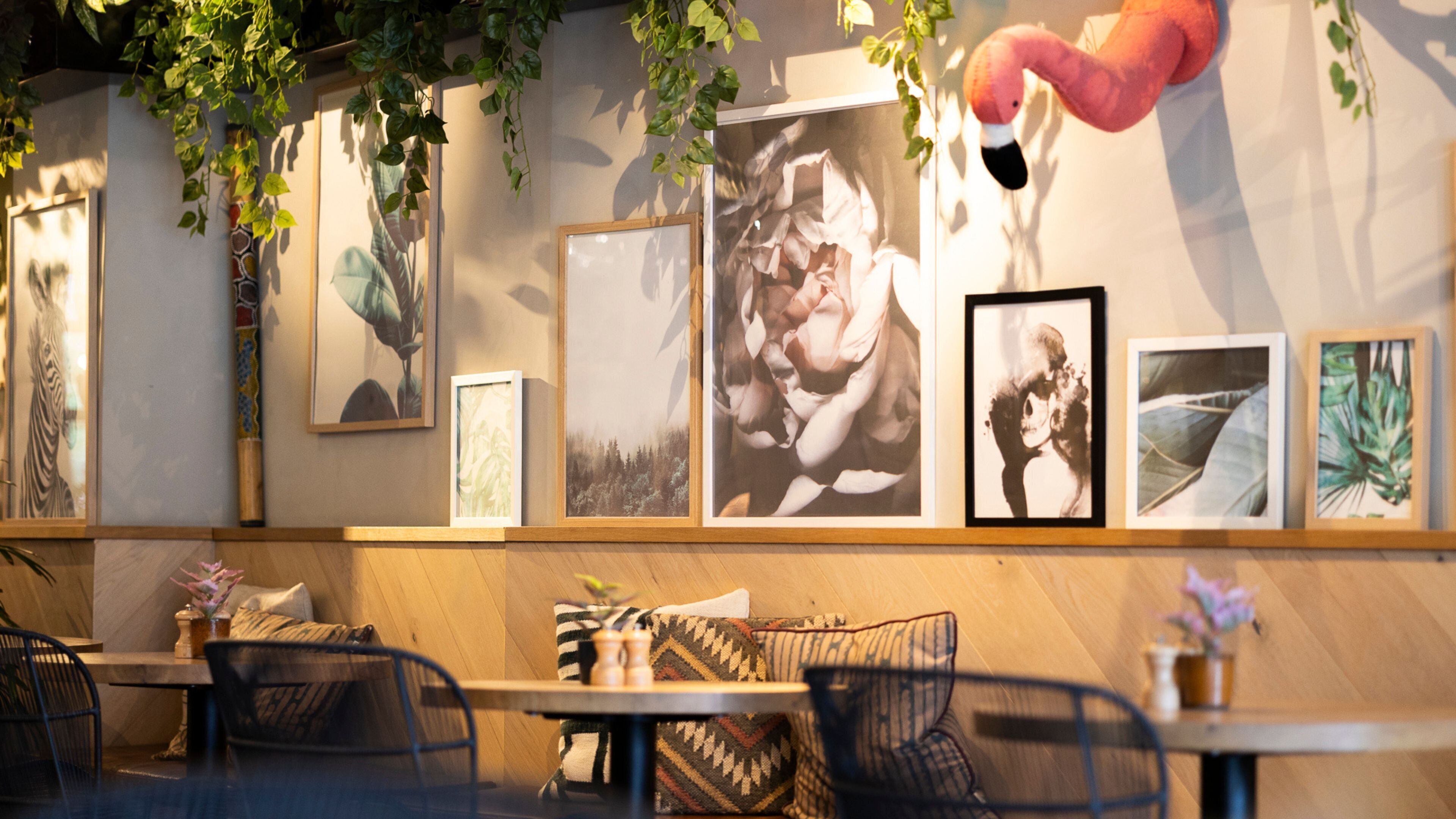 Interior de restaurante con mesas y sillas de madera, pared decorada con cuadros y plantas colgantes.