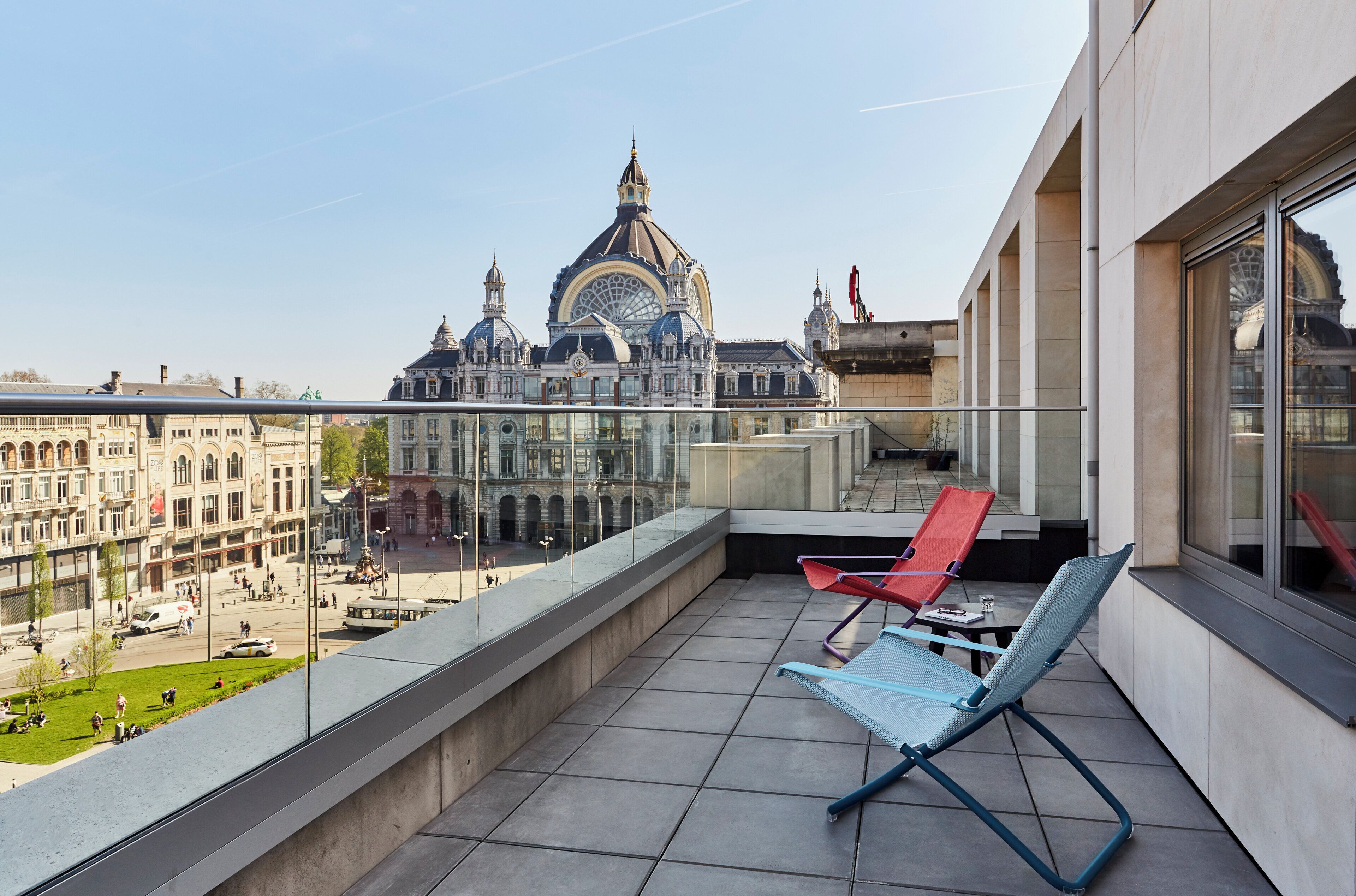 Een balkon met twee loungestoelen, een helder uitzicht op een historisch gebouw met koepels, en een tram op de achtergrond.