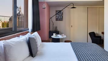 Une chambre d’hôtel comprenant un lit, un bureau, une chaise, une fenêtre donnant sur un immeuble et un luminaire suspendu.
