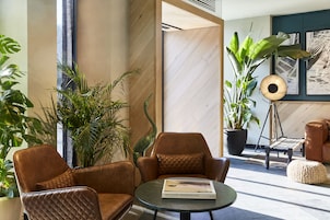 Uma área de estar moderna com poltronas de couro, uma mesa redonda com um livro e grandes vasos de plantas.