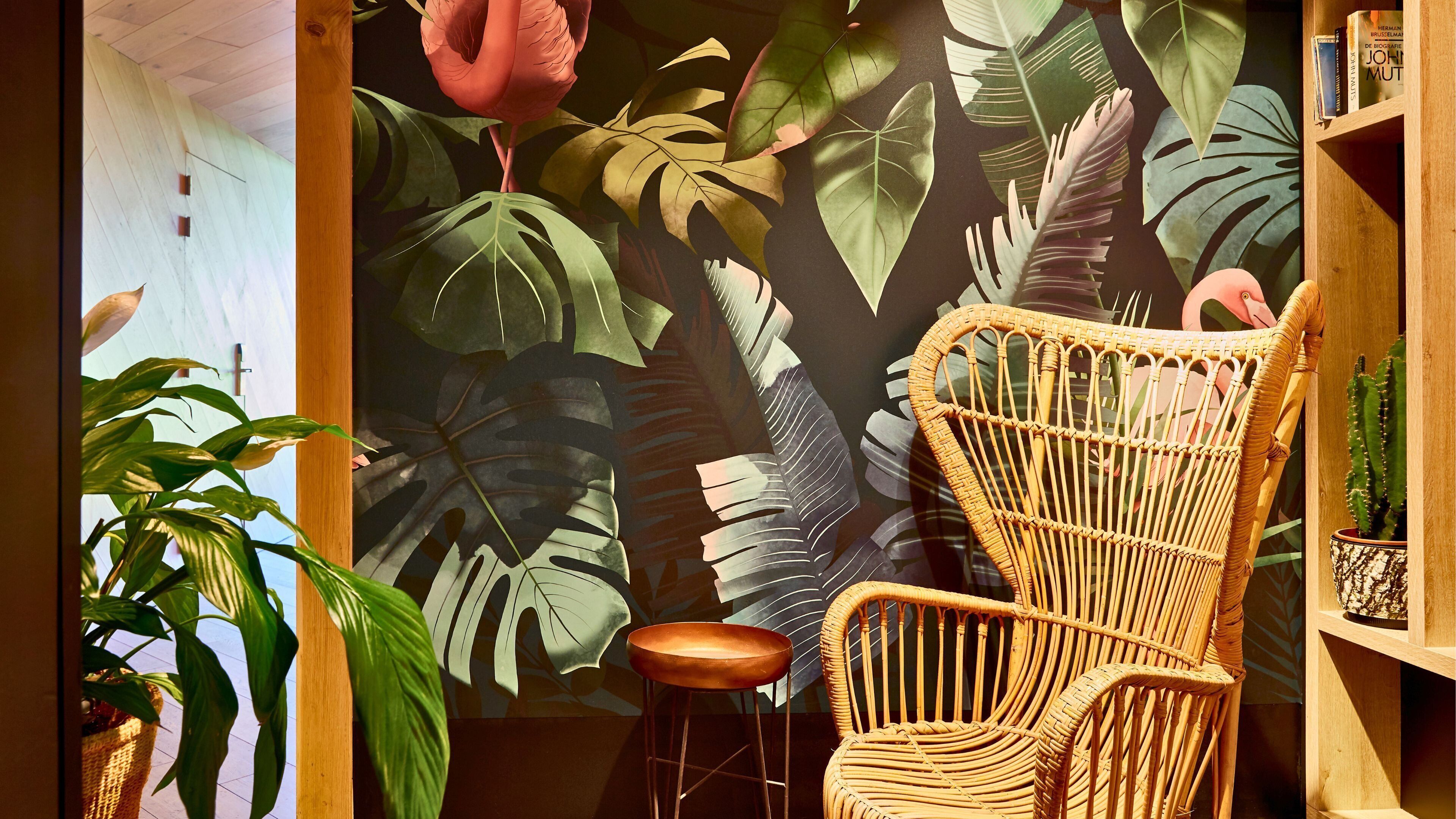 A room with a large tropical mural, a wicker chair, a wooden stool, and a bookshelf with books.