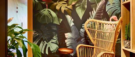 A room with a large tropical mural, a wicker chair, a wooden stool, and a bookshelf with books.