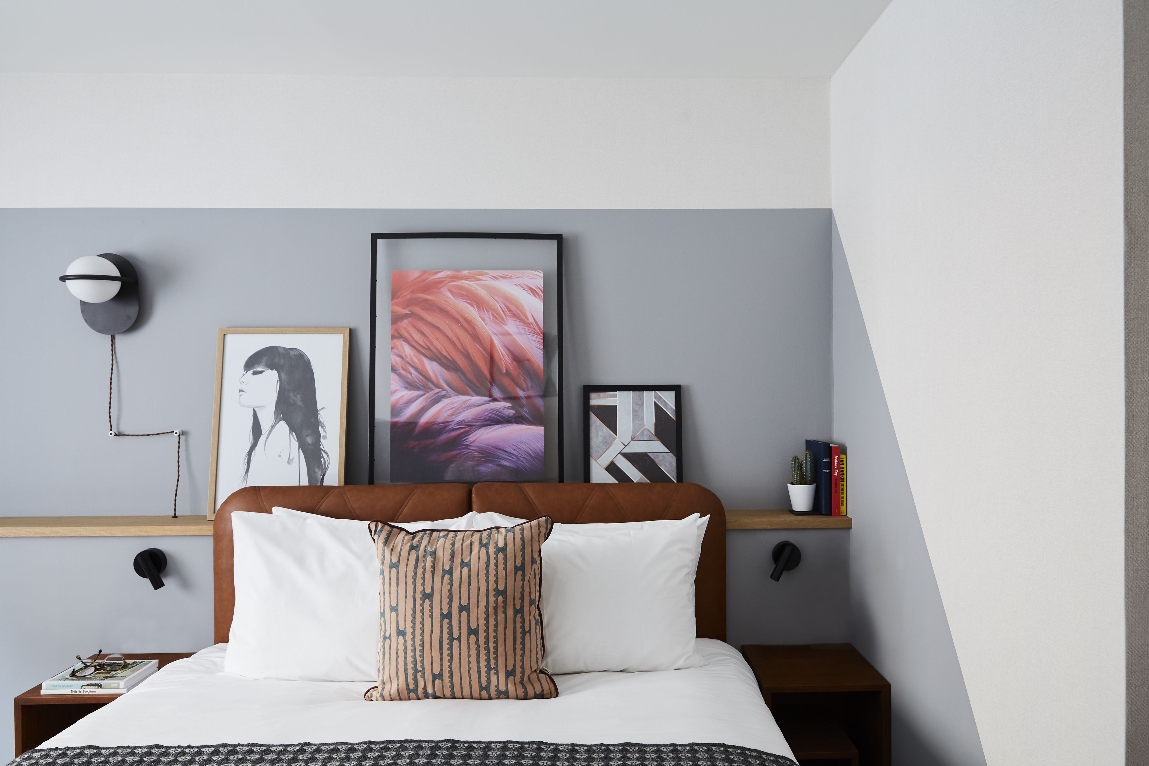 Een net opgemaakt bed met een gestreept kussen, een lijst met kunstwerk erboven, en een nachtkastje met boeken en een lamp.