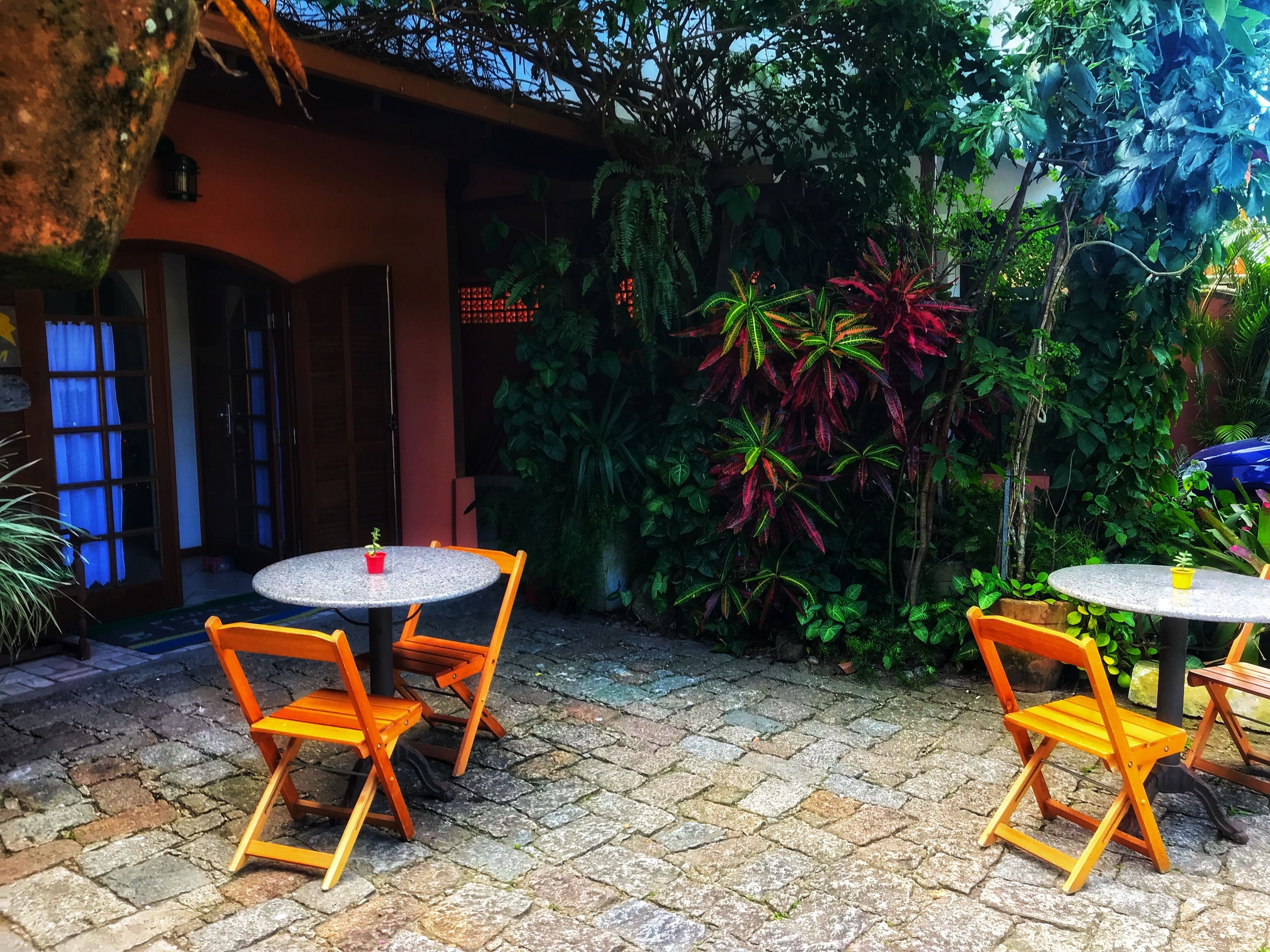 Un patio al aire libre con dos mesas redondas y cuatro sillas de madera, rodeado de abundante follaje y un suelo de piedra.