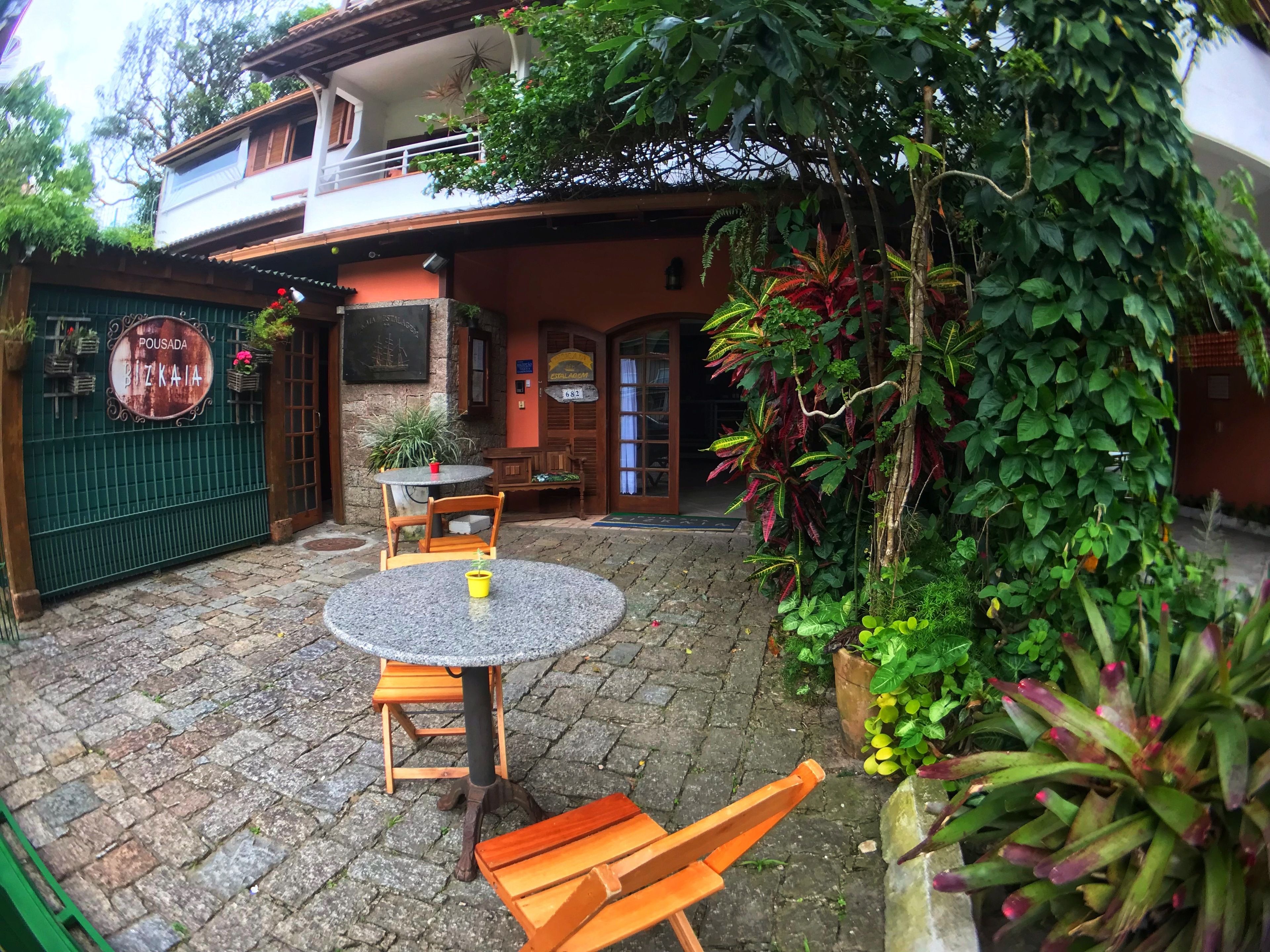 A courtyard with a round table and two wooden chairs, surrounded by potted plants and a green fence.