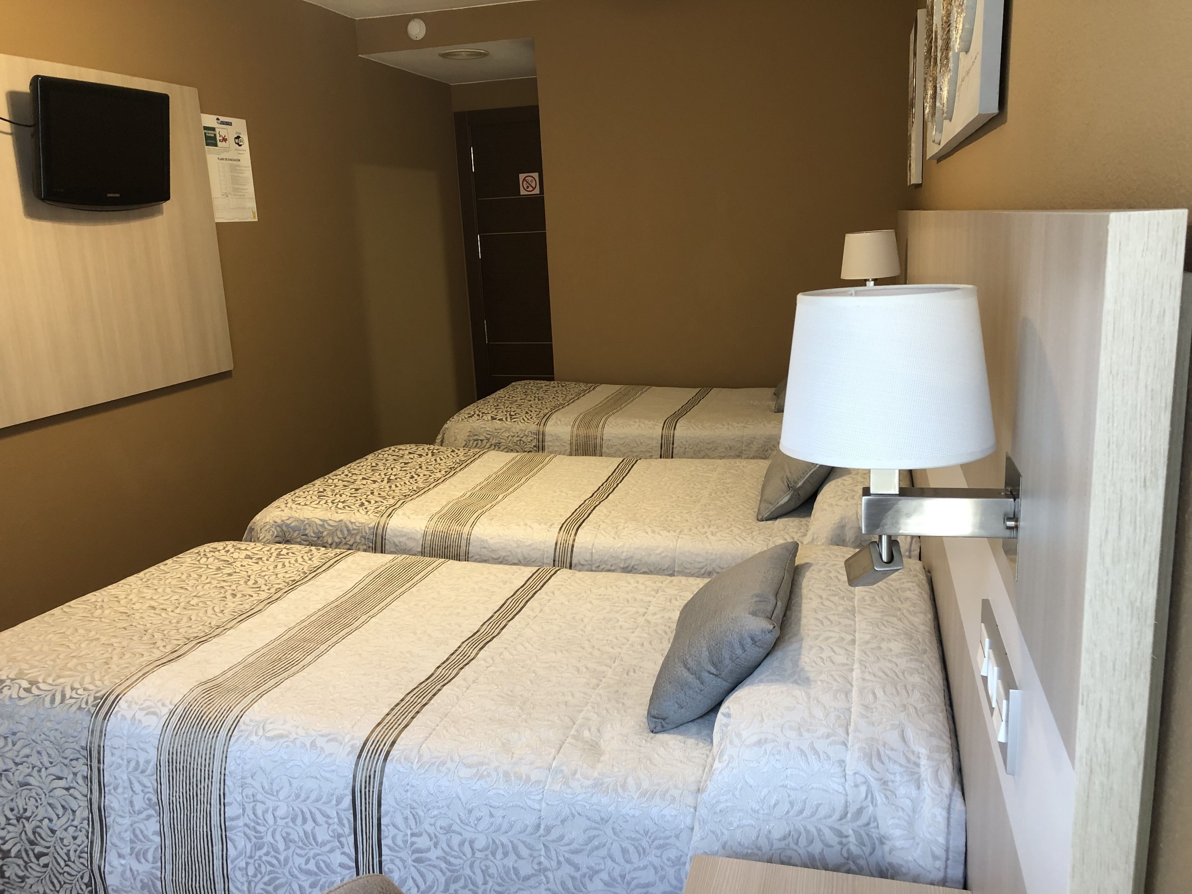 Habitación de hotel con dos camas, televisión montada en la pared, una lámpara y una puerta con un cartel.