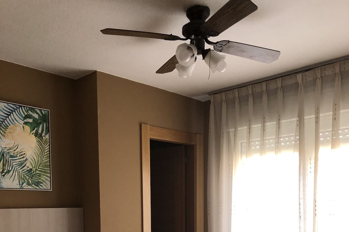 Um quarto com cama, ventilador de teto, mesa de cabeceira, telefone, porta e janela com cortinas.