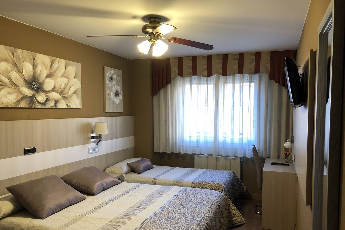 Quarto de hotel com duas camas, um ventilador de teto, uma grande pintura de flores, uma janela com cortinas, uma escrivaninha com abajur e uma TV fixada na parede.