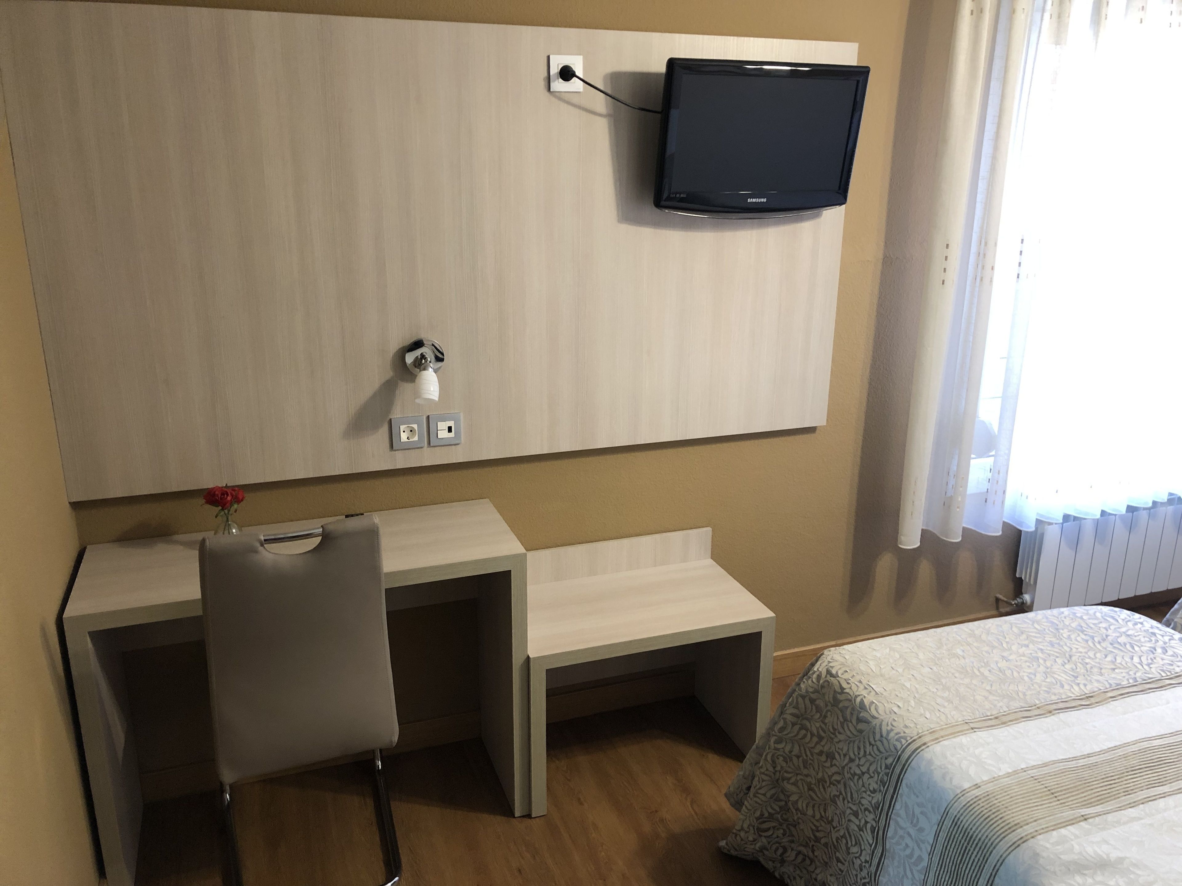 Habitación de hotel con cama, escritorio con silla, televisor en la pared y ventana con cortinas.