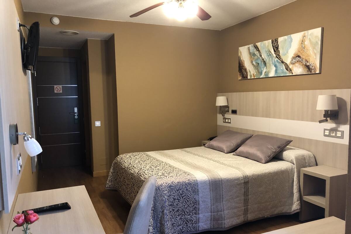 Quarto de hotel com cama, mesas de cabeceira, escrivaninha, cadeira, um vaso com flores, um quadro na parede e um ventilador de teto.