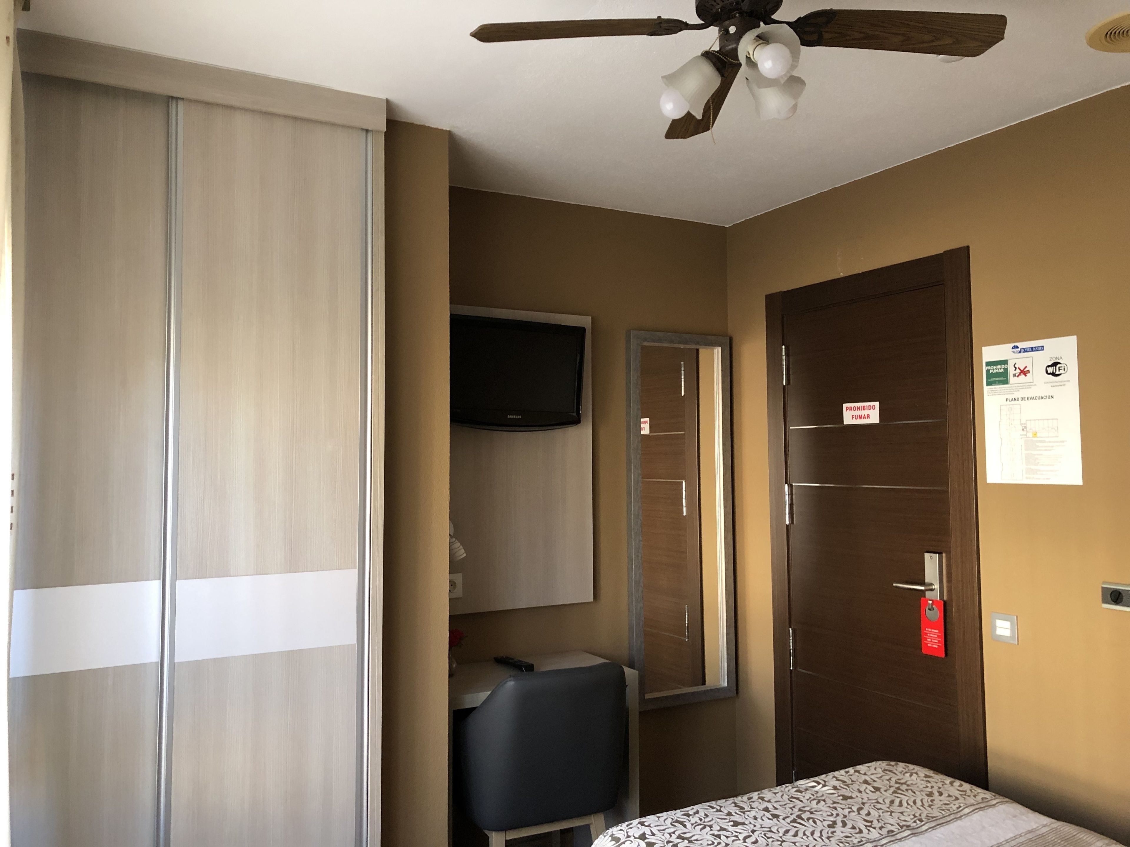 Habitación de hotel con cama, armario, escritorio, silla, televisor, ventilador de techo y puerta con señal de seguridad contra incendios.