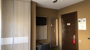Habitación de hotel con cama, armario, escritorio, silla, televisor, ventilador de techo y puerta con señal de seguridad contra incendios.
