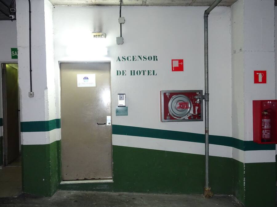 Ein Flur mit einer grün-weiß gestreiften Wand, einem Rauchmelder und einer Tür mit der Aufschrift 'ASCENSOR DE HOTEL'.