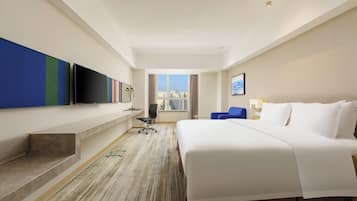 Ein Hotelzimmer mit einem großen Bett, einem flachbildfernseher an der Wand, einem Schreibtisch mit Stuhl und einem Fenster mit Blick auf die Stadt.