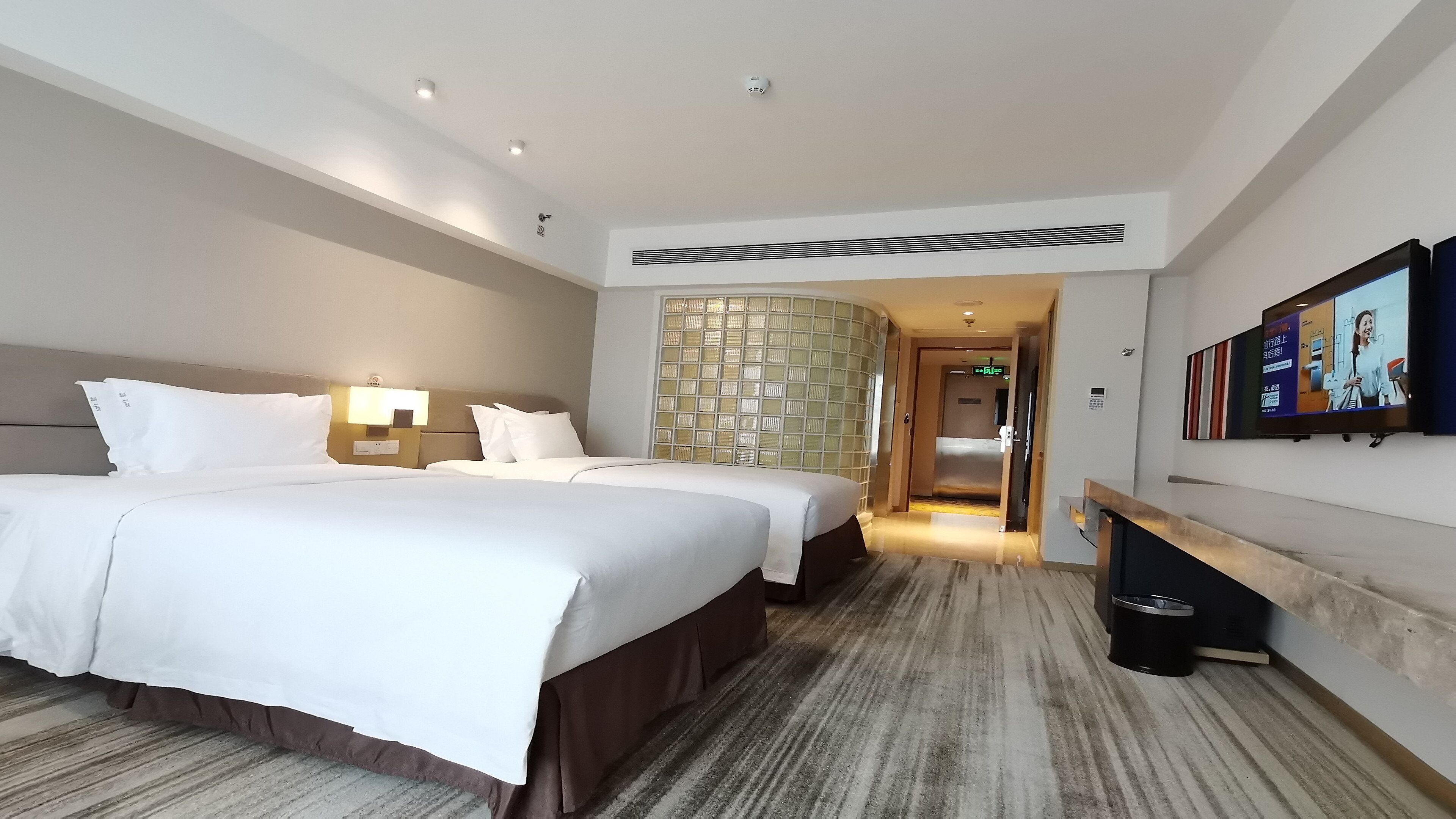 A hotel room with two beds, a TV, a wall-mounted heater, and a reception area visible through an open door.